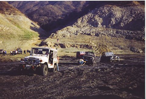 1992 Ford Bronco - Several off-road vehicles are stuck in thick mud in a dry, hilly area, with people gathered around.