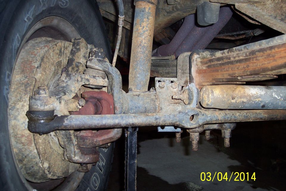 Rusty front knuckle-joint with ball joint, tie rod end, and leaf spring assembly. Tire is visible on the left