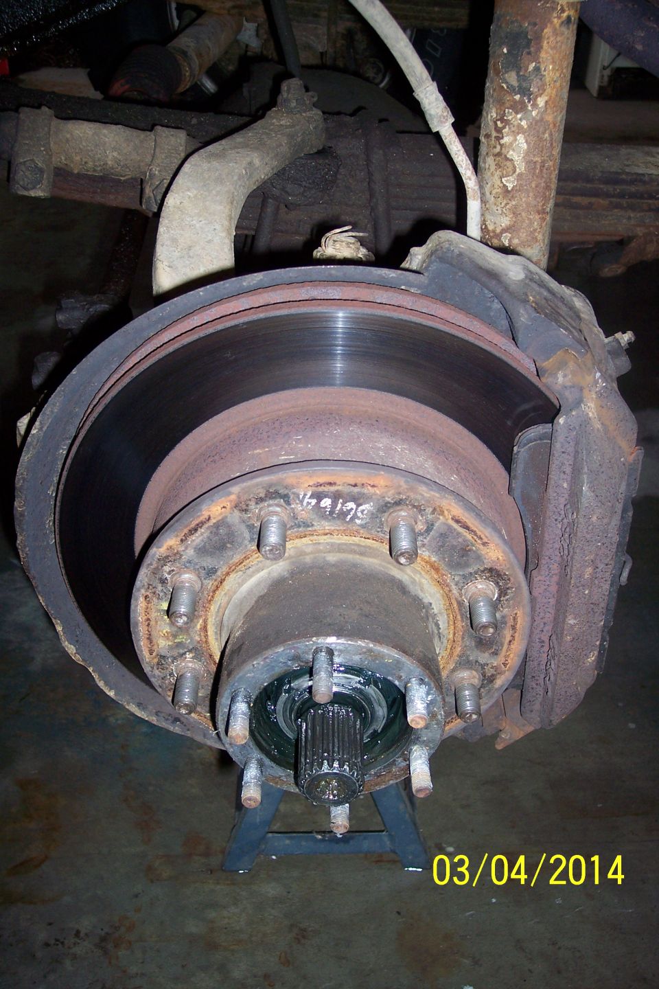 Rusty front wheel hub with brake rotor and caliper, supported by a jack stand. White writing on rotor reads '46129'