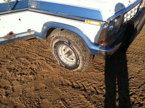 1979 Ford Bronco - Front corner of a 1979 Ford Bronco, showing its blue fender flares and chrome bumper, partially obscured by dirt and tire tracks.