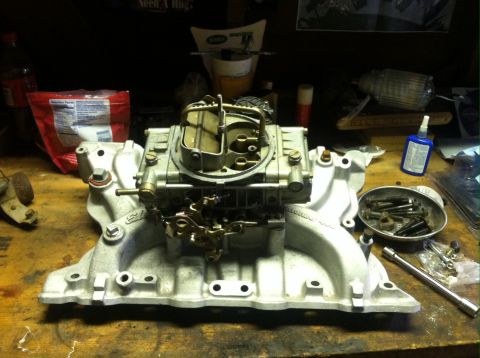 1979 Ford Bronco - A carburetor and intake manifold sit on a cluttered workbench, surrounded by tools and other items.