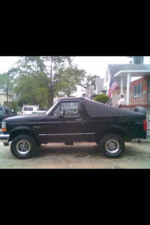 1995 Ford Bronco - This black Ford Bronco is parked on a residential street with a house and trees in the background.