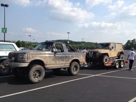 1995 Ford Bronco - A muddy 1995 Ford Bronco is seen towing a Jeep on a trailer in a parking lot.