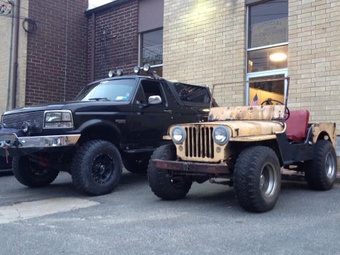 1995 Ford Bronco - A black 1995 Ford Bronco is parked next to a weathered yellow Jeep in front of a brick building.