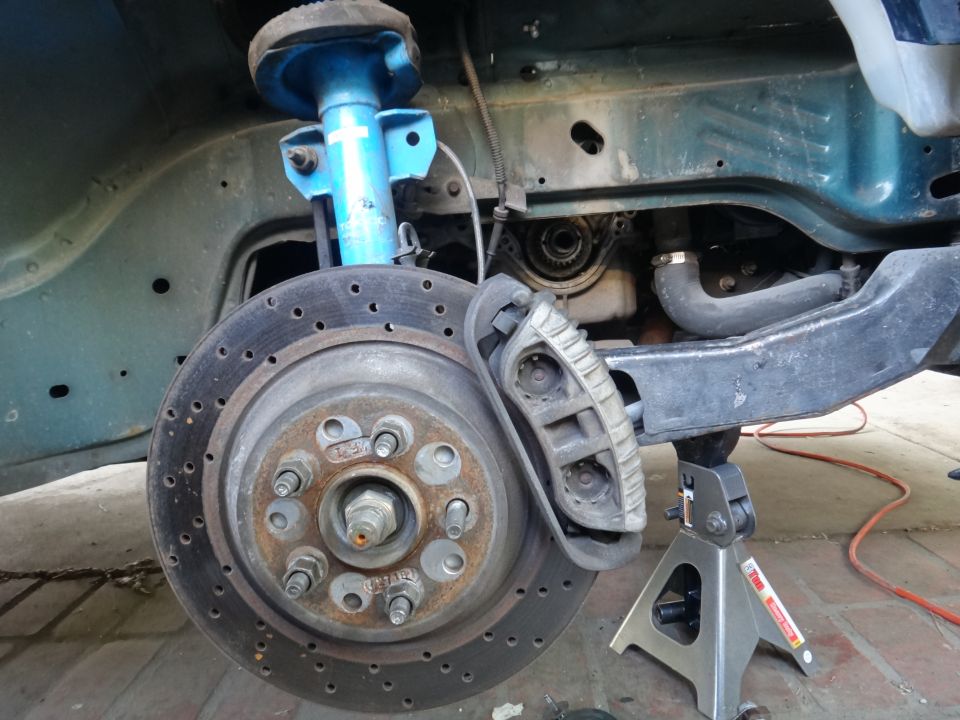 Close-up of a car's front brake rotor, caliper, and blue shock absorber, with engine components visible behind