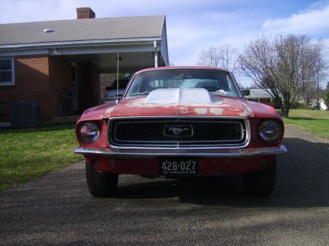 1968 Ford Mustang - A red 1968 Ford Mustang sits in a driveway, its front end showing signs of wear and tear, including a prominent hood scoop.