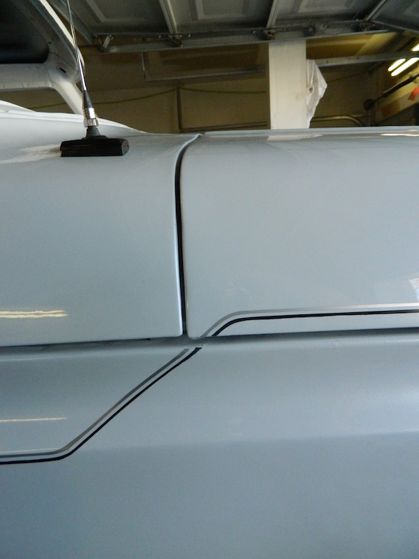Close-up of a light-colored vehicle's side panel showing a seam and pinstripe detail. An antenna is visible on top