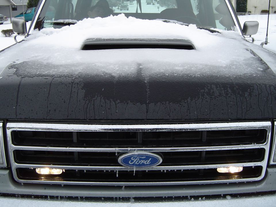 Snow-dusted black cowl-induction hood and chrome grille of a Ford with blue oval