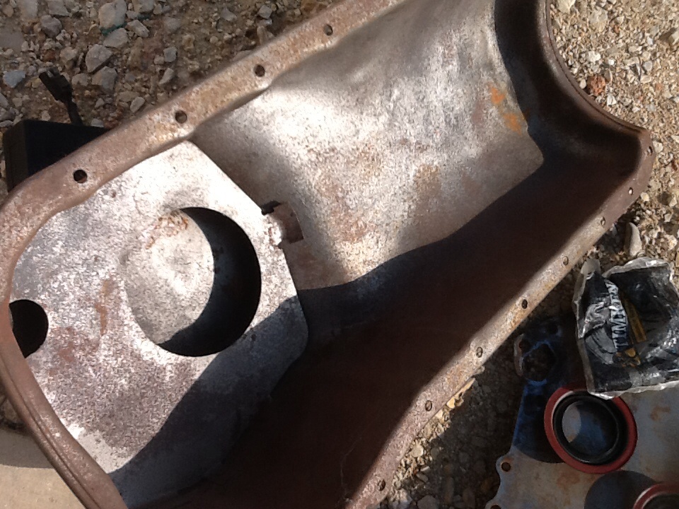 Rusty metal oil pan with circular cutout and bolt holes, next to engine parts