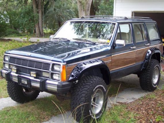 1984 Jeep Cherokee XJ with wood paneling and off-road tires parked outside a garage