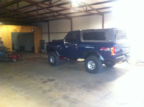 1982 Ford Bronco - A dark blue Ford Bronco with a soft top is parked inside a garage, showing its '4x4' badging.