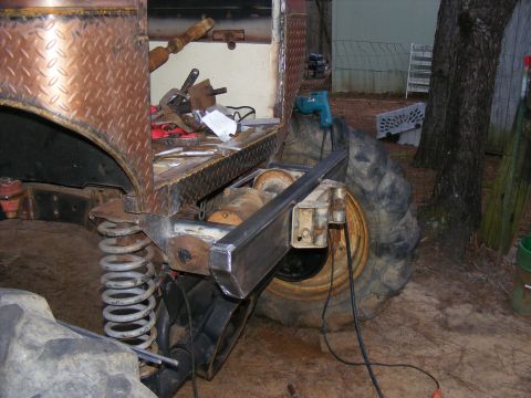 Custom off-road vehicle rear axle, large tire, and suspension components. Diamond plate bodywork visible