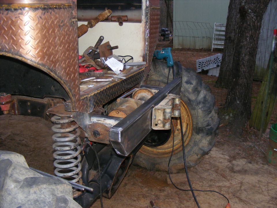 Custom off-road vehicle rear axle, large tire, and suspension components. Diamond plate bodywork visible