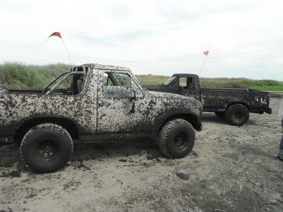 Two mud-covered trucks, one a Bronco-style, parked on a dirt track with grassy dunes behind them