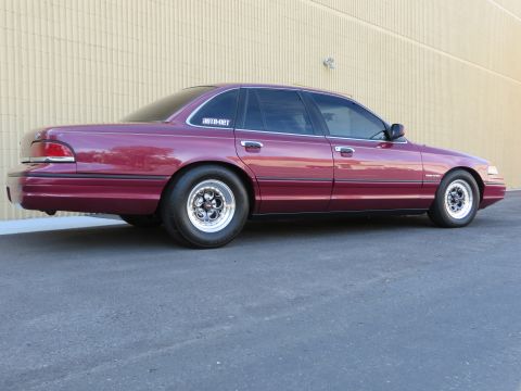 1992 Ford Crown Victoria - Side rear view of a maroon Ford Crown Victoria sedan with chrome wheels parked beside a tan wall.