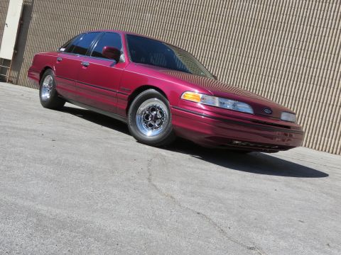 1992 Ford Crown Victoria - Front three-quarter view of a maroon Ford Crown Victoria sedan with chrome wheels on pavement against a corrugated wall.