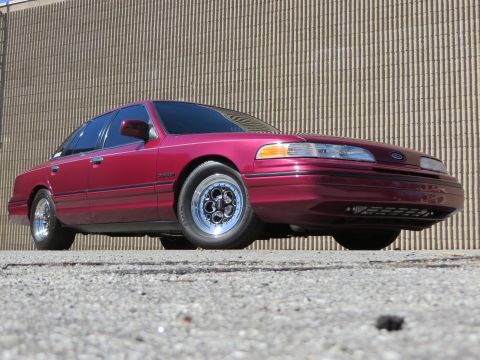 1992 Ford Crown Victoria - A maroon 1992 Ford Crown Victoria is shown from a low angle, highlighting its custom wheels against a neutral background.