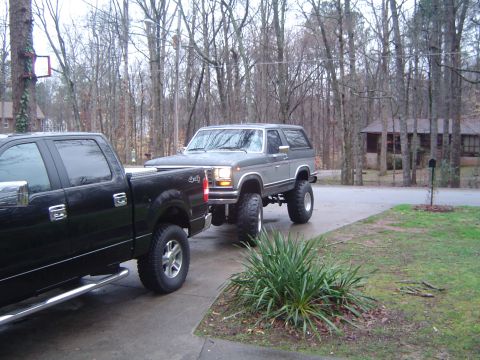 1986 Ford Bronco - A lifted gray Ford Bronco is parked on a driveway next to a black pickup truck.