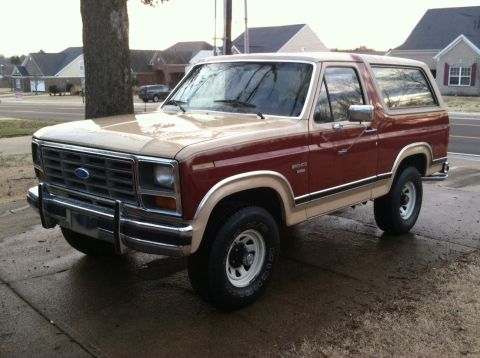 1986 Ford Bronco - A two-tone red and tan Ford Bronco is parked on a wet driveway with houses in the background.