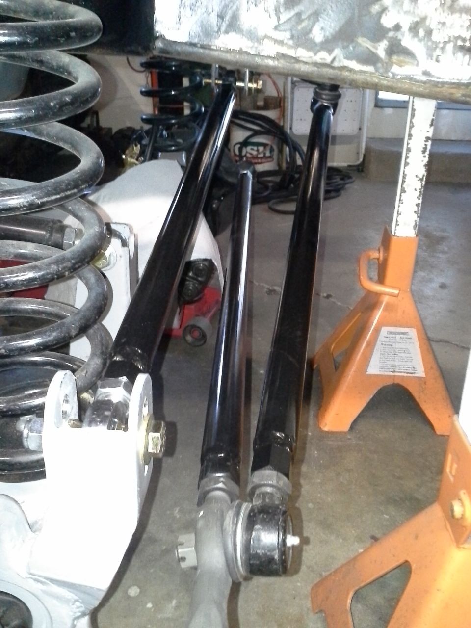 Vehicle suspension components, including control arms and coil springs, supported by orange jack stands in a garage