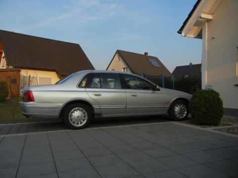 1999 Ford Fairlane - A silver 1999 Ford Fairlane sedan is parked on a paved driveway in front of houses.