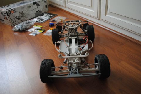 1986 Ford Bronco - The chassis of a remote control car, with suspension and electronic components visible, sits on a wooden floor next to its box.