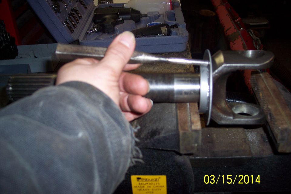 Hand holding a tool against a metal shaft clamped in a vise, with a u-joint visible