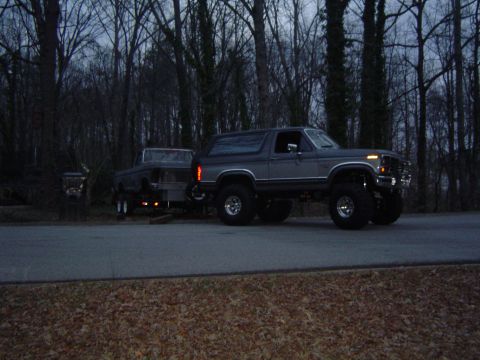 1986 Ford Bronco - A lifted gray Ford Bronco tows a trailer on a road at dusk, with a line of trees behind it.