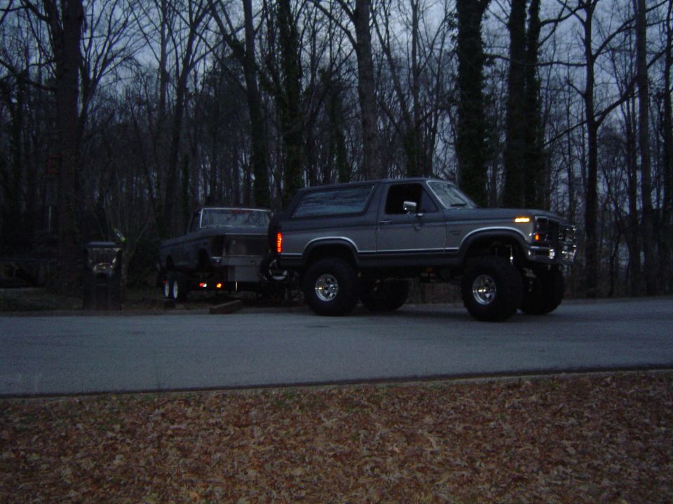 Gray lifted Ford Bronco towing a dark trailer on a road with trees in the background