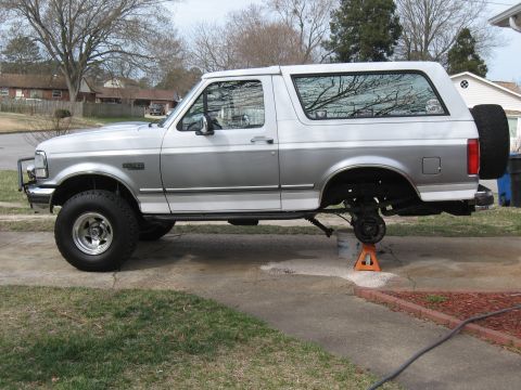 1994 Ford Bronco - A silver 1994 Ford Bronco is shown with its rear wheel removed and the vehicle supported by jack stands.