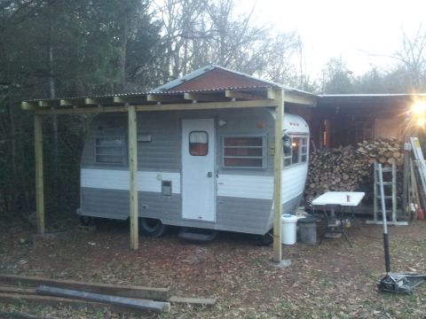 1986 Ford Bronco - A travel trailer is parked outdoors under a makeshift awning, with firewood stacked to the right.