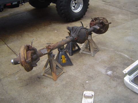 1986 Ford Bronco - Front axle assembly resting on jack stands, with a portion of a large tire and wheel visible in the background.