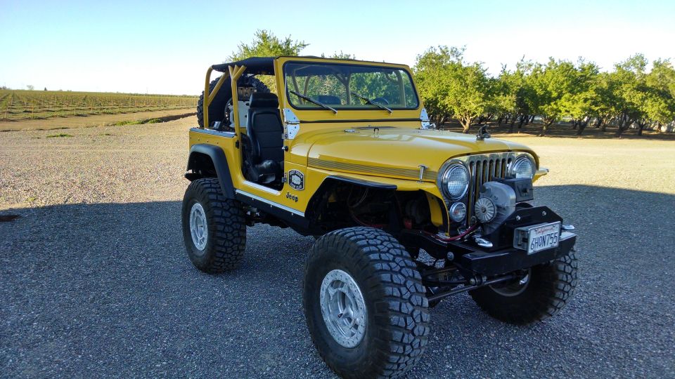 Yellow Jeep CJ with large tires and a winch on the front bumper. It's parked on a gravel surface with trees and a field in the background