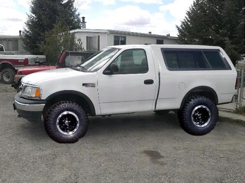 1994 Ford Bronco - A white pickup truck with a camper shell and oversized tires is parked outdoors, with other vehicles visible in the background.