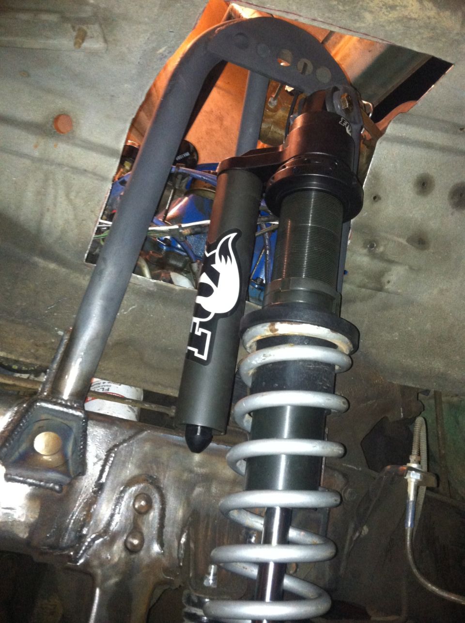 Close-up of a coilover suspension setup with a black Fox shock absorber and silver spring, mounted to custom metalwork