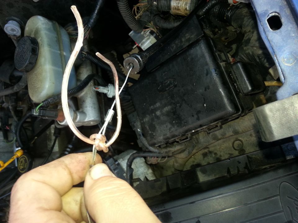 Close-up of a blue vehicle's engine bay with a hand holding wires near a black fuse box and brake fluid reservoir