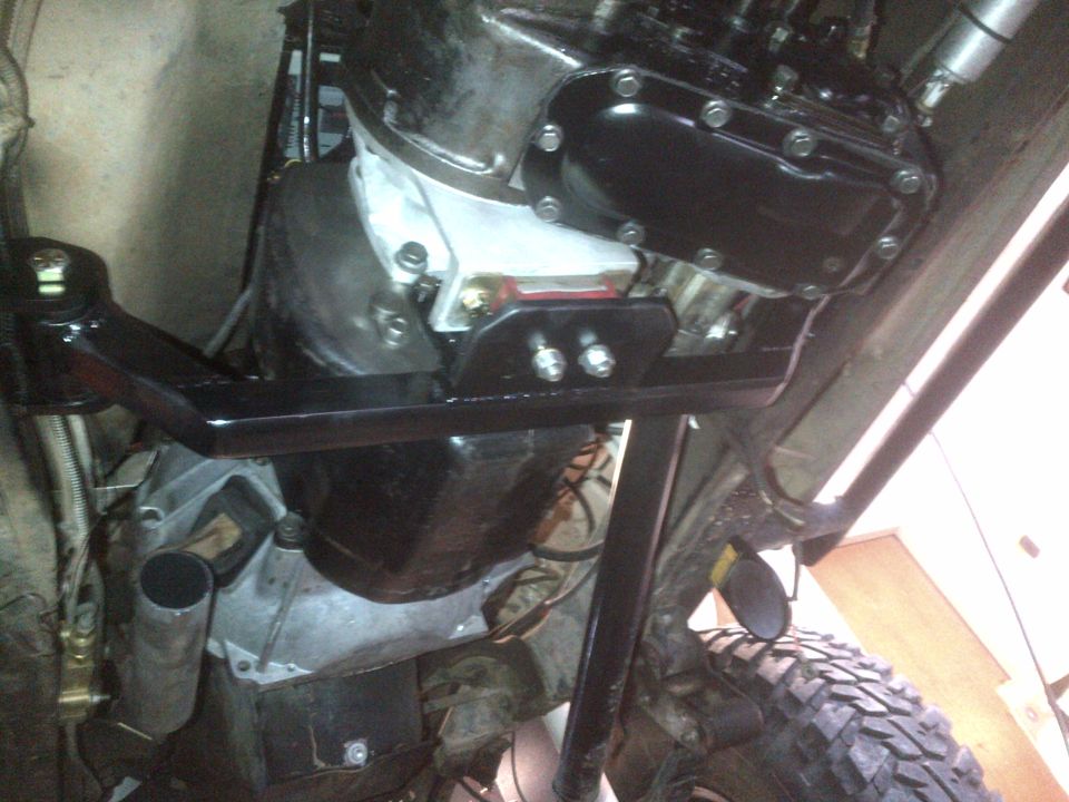 Underside view of a Jeep CJ drivetrain with a black transmission housing and silver transfer case, mounted with a black bracket