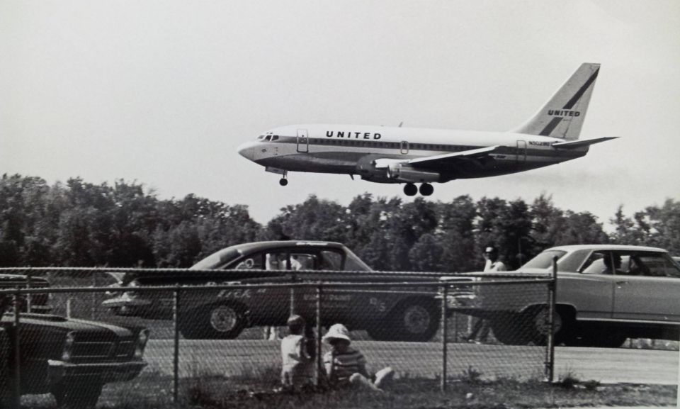 United Airlines jetliner descending over cars and spectators