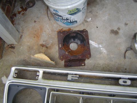 1986 Ford Bronco - Vehicle parts, including a headlight bucket and grille, lie on a concrete surface next to a bucket of detergent.
