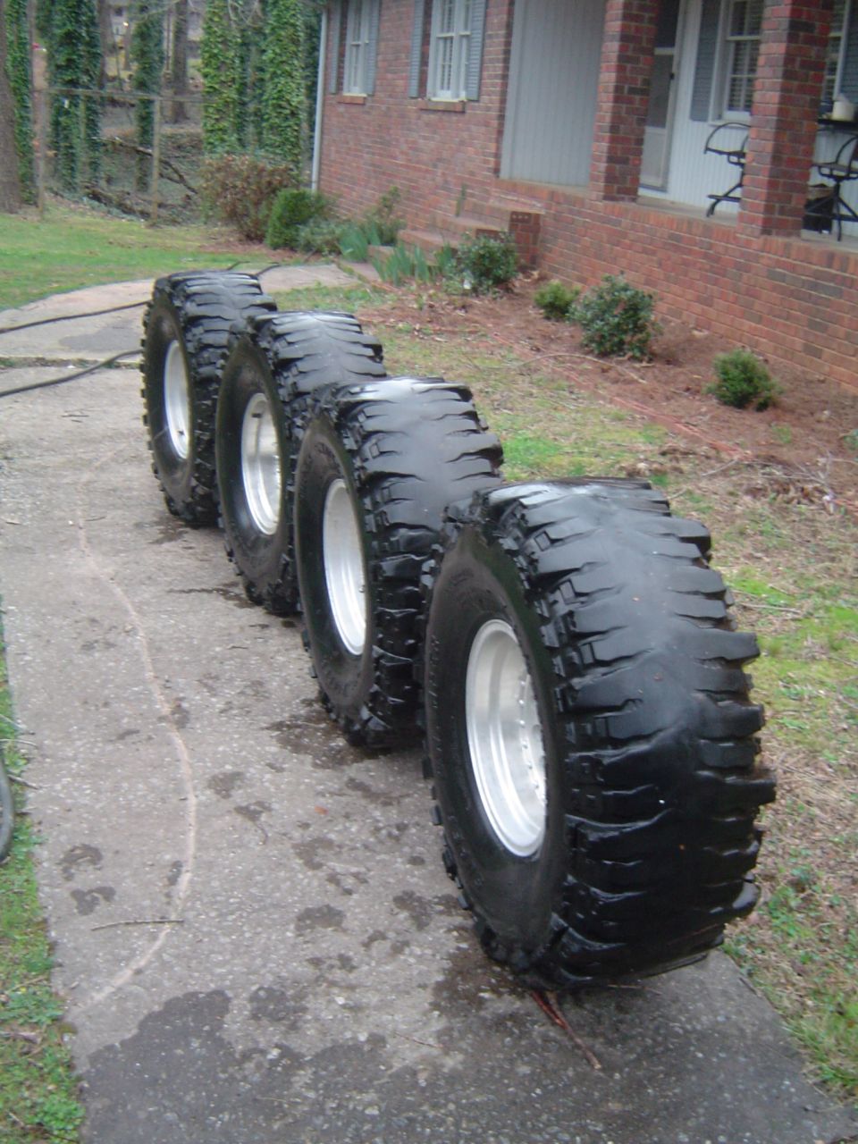 Four large off-road tires lined up on a concrete path in front of a brick house