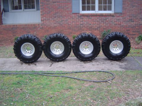 1986 Ford Bronco - Four off-road tires and rims are displayed on a concrete walkway in front of a residential building.