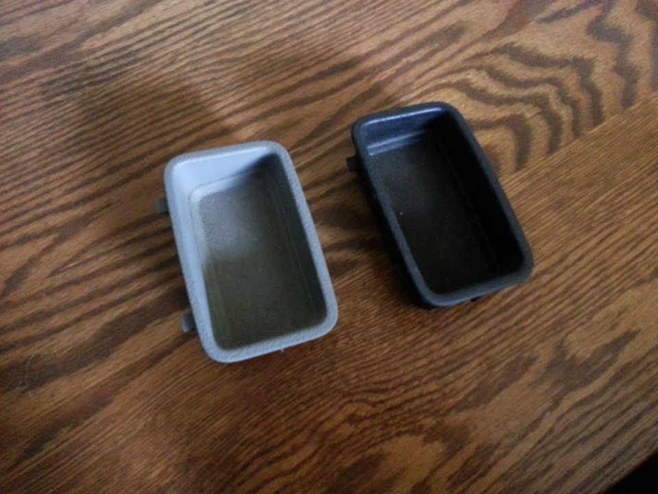 Two rectangular plastic car interior trim pieces, one gray and one black, on a wooden surface