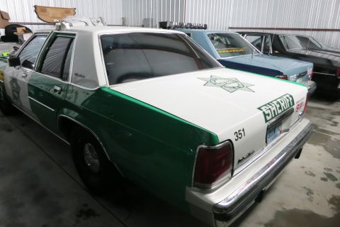 1991 Ford LTD Crown Victoria - A sheriff's car, marked with a star emblem and the word SHERIFF, is parked in a garage with other vehicles.