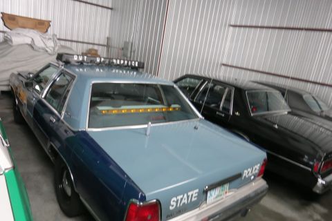 1991 Ford LTD Crown Victoria - A light blue police car with an "OUT OF SERVICE" light bar is parked inside a garage next to other vehicles.