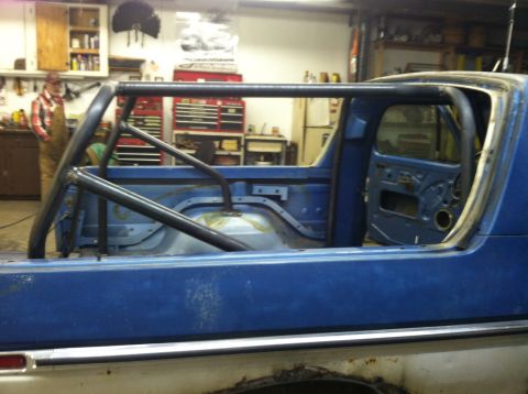 1979 Ford Bronco - The interior of a blue Ford Bronco is visible, showing a custom roll cage and removed door panel. A person stands in the background of a wo…