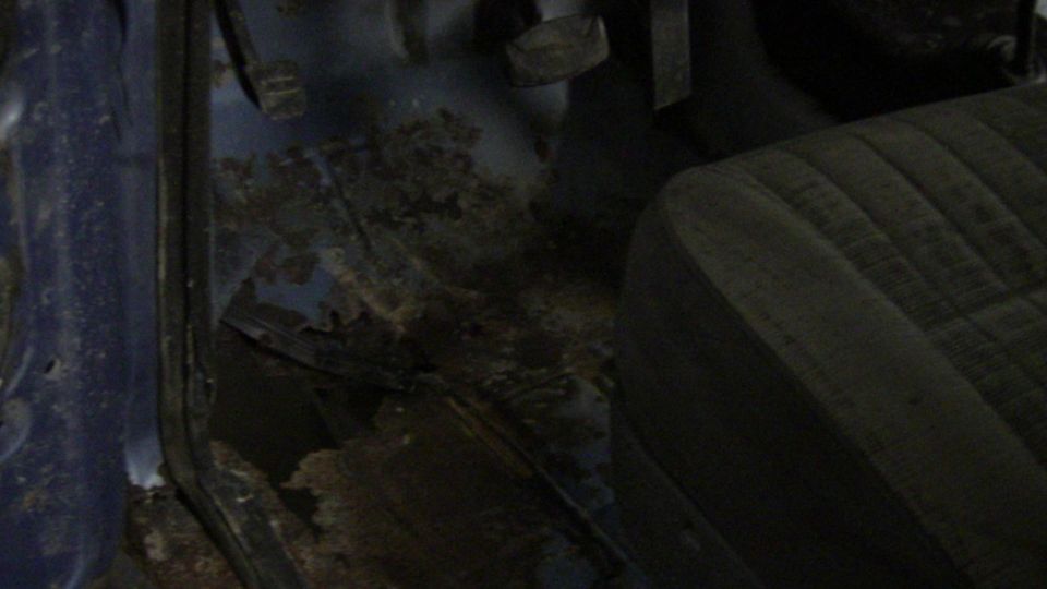 Interior floor of a vehicle showing significant rust damage and a portion of the driver's seat