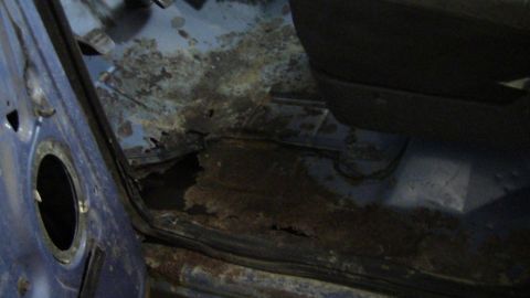 1979 Ford Bronco - The interior floor of a blue vehicle shows significant rust damage and holes, with a portion of the seat visible.