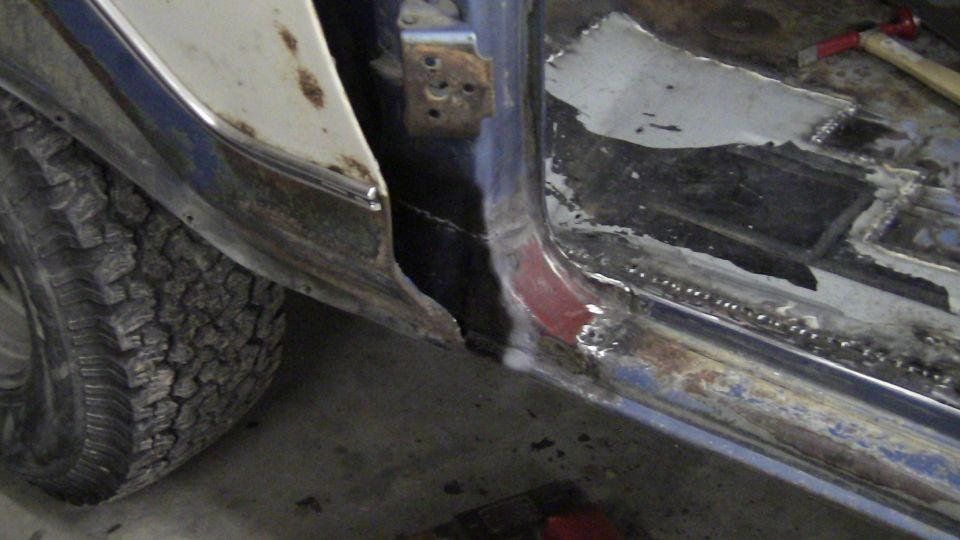 Vehicle door frame with rust and repair work, showing a tire and part of the body. The metal is exposed and welded
