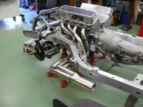 Car chassis on red stands with V8 engine, chrome headers, and exhaust pipes. Some boxes are stacked near the f