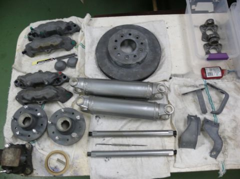 Car parts including brake calipers, rotors, driveshafts, and hubs laid out on a white cloth. A bottle of red L